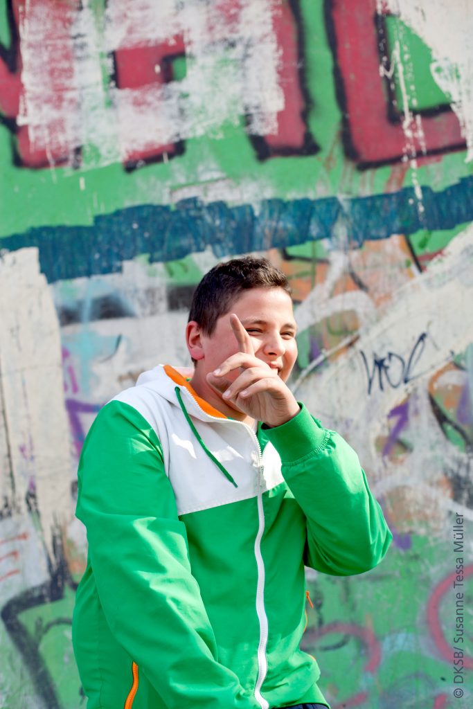 Junge in grüner Jacke, der vor einer graffitibedeckten Wand steht und mit einer Hand gestikuliert.