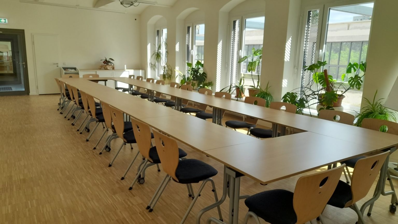 Foto eines langen Konferenz- oder Besprechungsraums mit zwei Tischreihen, die in U-Form angeordnet sind, und mehreren Holzstühlen mit schwarzen Polstern. Große Fenster an einer Seite lassen Tageslicht in den Raum strömen, und mehrere Topfpflanzen stehen auf den Fensterbänken und Tischen und schaffen eine helle und einladende Atmosphäre.