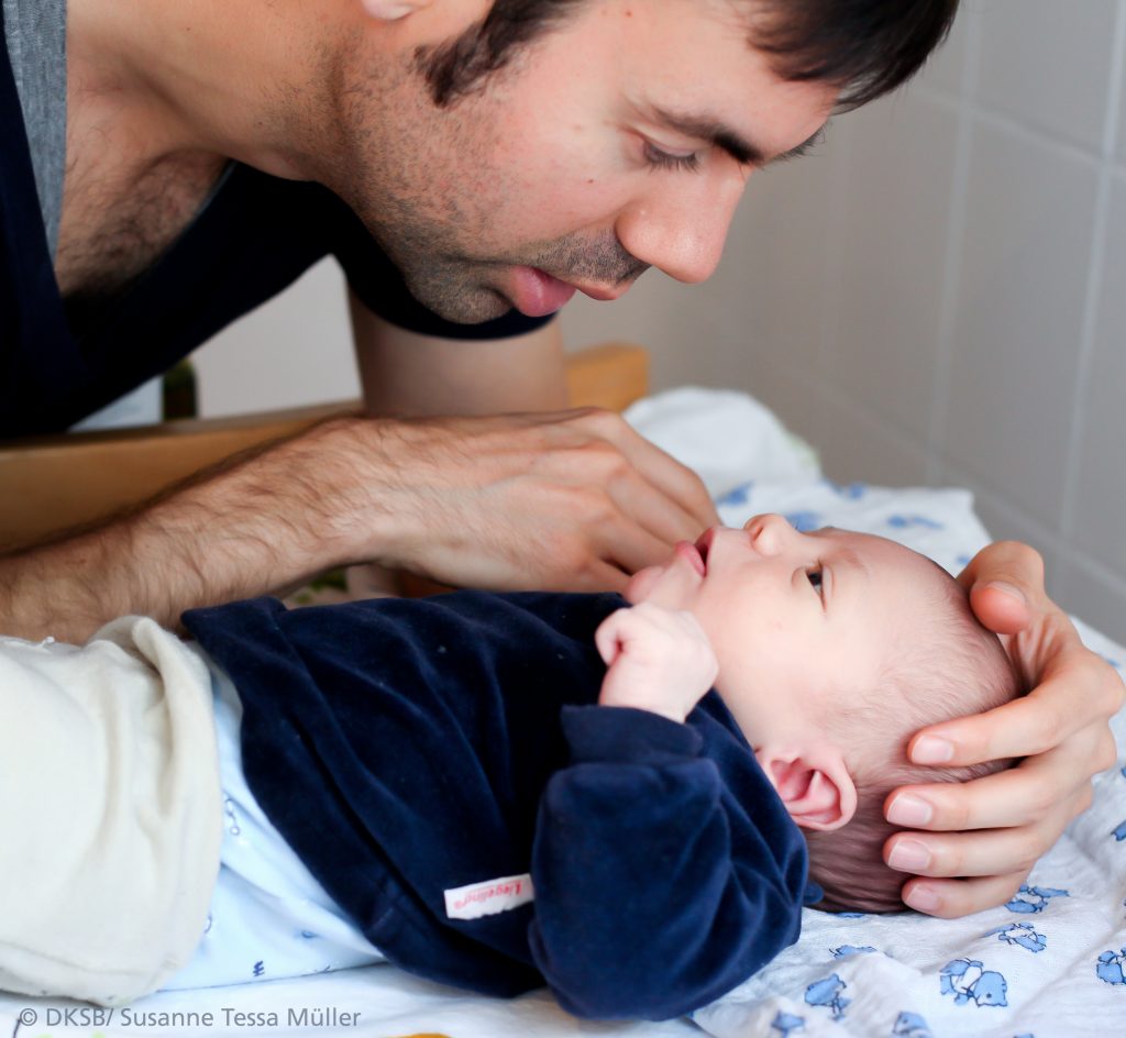 Ein Vater, der mit seinem Baby auf einer Decke interagiert und es sanft anfasst.