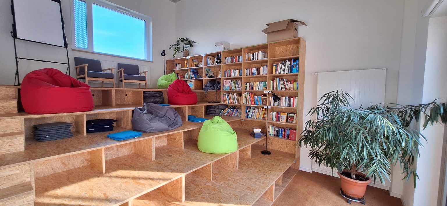 Foto eines modernen Lese- oder Entspannungsbereichs mit gestuften Holzsitzgelegenheiten und bunten Sitzsäcken in Rot, Grau und Grün. Der Raum verfügt über ein großes Bücherregal voller Bücher, eine Topfpflanze und natürliches Licht, das durch ein Fenster fällt und eine gemütliche und einladende Atmosphäre schafft.