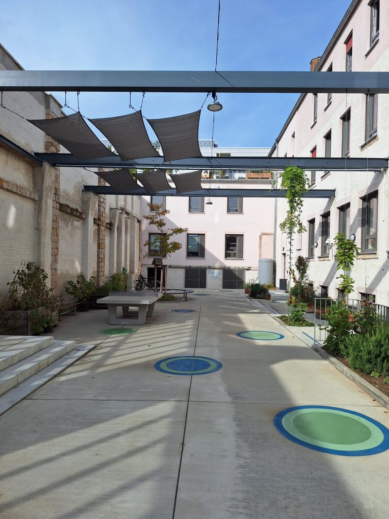 Innenhof mit Sonnenschutzsegeln und bunten Kreisen auf dem Boden in einem urbanen Umfeld.