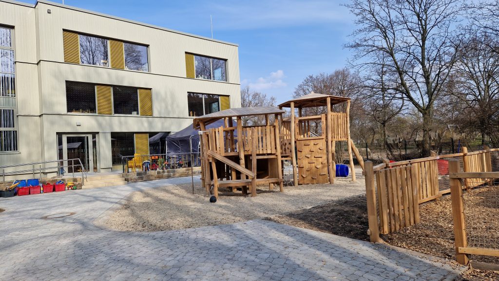 Außenbereich des Kinderhaus „Regenbogen“ in Dresden: hölzernes Spielgerät, gelbe Akzente am modernen Gebäude, gepflasterter Hof und Bäume im Hintergrund.