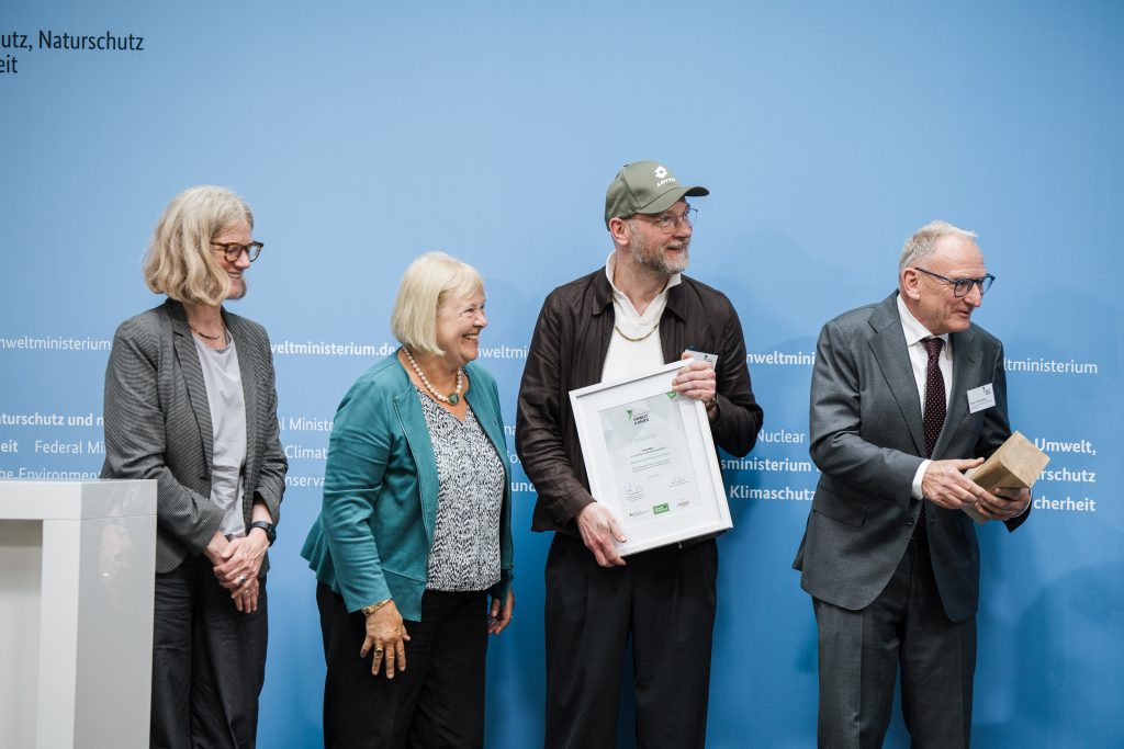 Vor blauem Hintergrund hält Alexander Pötzsch Architekturen die Urkunde im Rahmen beim Bundespreis Umwelt & Bauen für den Kinderschutzbund Ortsverband Dresden e.V.