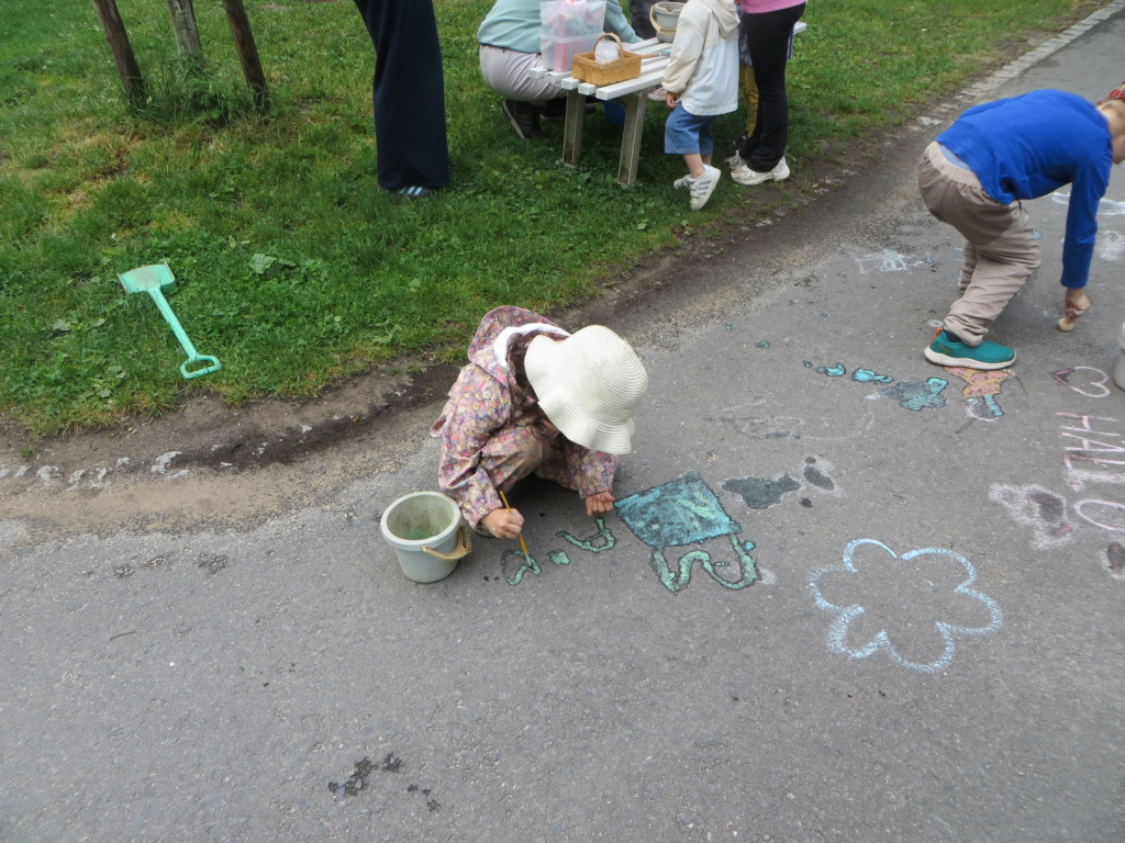 Kinder malen beim Kindertag mit Kreide bunte Wasserbilder und Figuren auf den Gartenweg, während ein Kind im weißen Hut kniet.