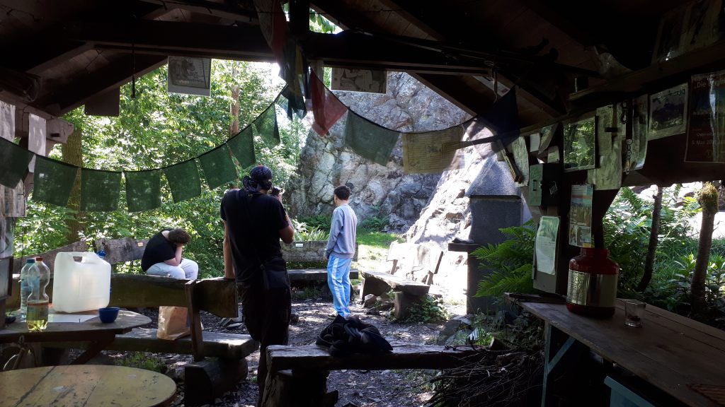 Drei Jugendliche stehen und sitzen in einer offenen Waldhütte am Fels beim Oderwitzer Spitzberg, während grüne Wimpel und Wegweiser hängen.